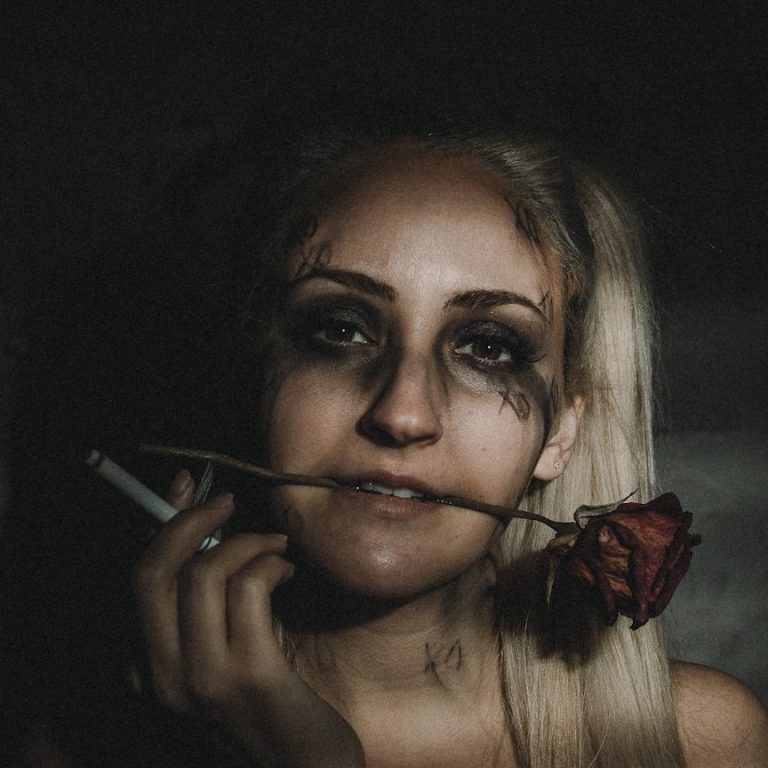 Frau mit rauchendem Blick, einer Rose im Mund und geschminktem Gesicht.