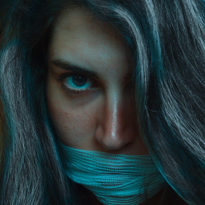 Frau mit langen, grauen Haaren und einem Band um den Mund, intensiv blickend.