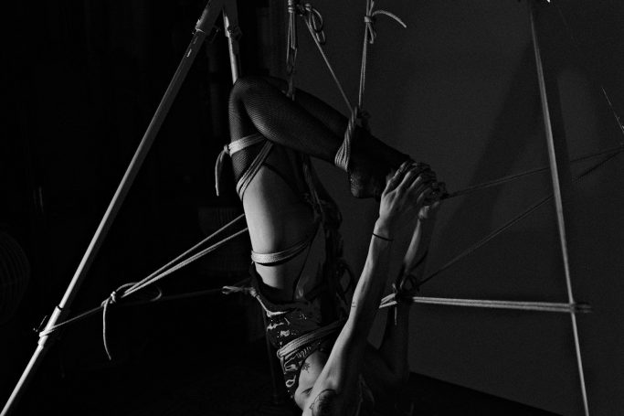 Person in a harness, suspended and performing acrobatic movements in a dark setting.
