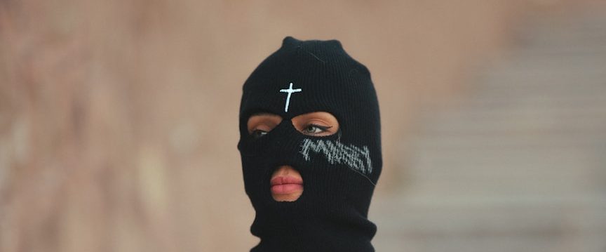 Person mit schwarzer Maske, gekennzeichnet durch ein Kreuz und Schriftzüge.